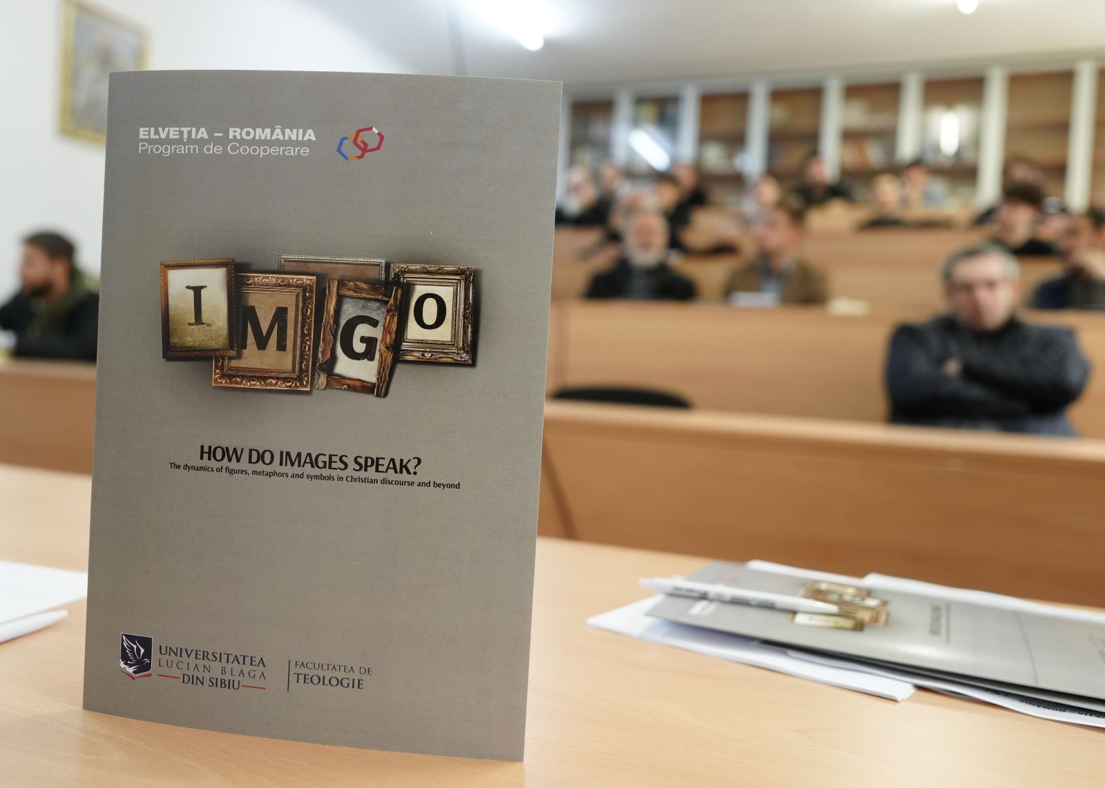 Flyer des Projekts im Vordergrund, im Hintergrund Auditorium mit Teilnehmenden des Events