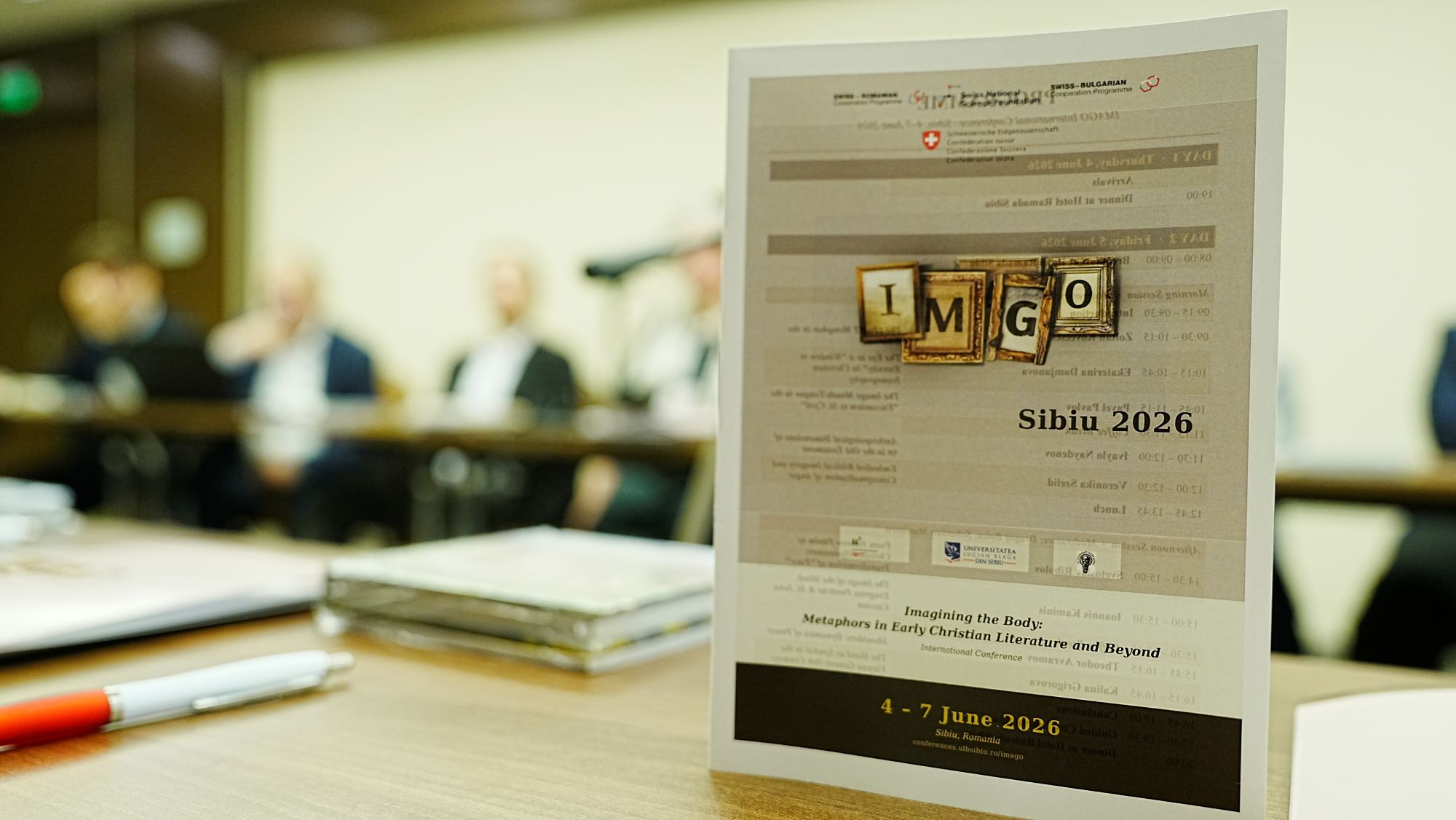 Flyer der Konferenz auf dem Tisch, im Hintergrund sieht man unscharf die Konferenzteilnehmer